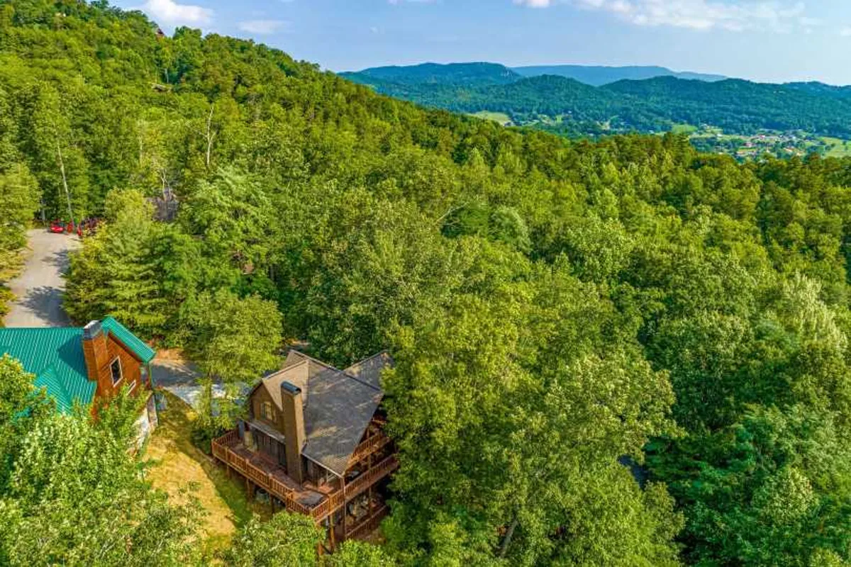 A charming wooden cabin nestled among tall trees with a breathtaking mountain view in the background. The peaceful setting is complemented by soft evening light, inviting guests to relax on the deck and soak in the natural beauty.