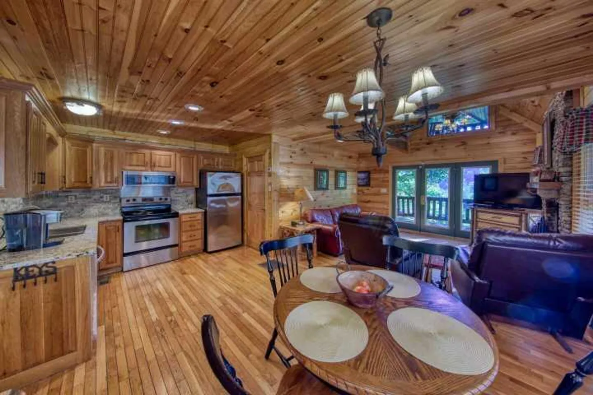 This image showcases a warm, welcoming interior featuring a round dining table, wooden cabinetry, a glowing chandelier, and a comfortable living area with large windows. Natural light fills the space, and the wooden accents highlight the cabin's rustic charm, making it an inviting spot for shared meals or intimate gatherings.