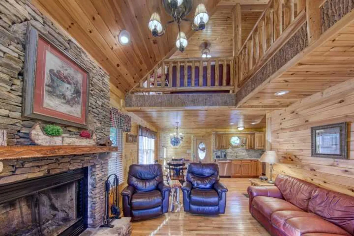 A spacious great room with a prominent stone fireplace as the focal point. The room features soaring timbered ceilings, rich wooden details, plush leather couches, and a dining area in the background. Warm, ambient lighting adds to the cozy, welcoming atmosphere.
