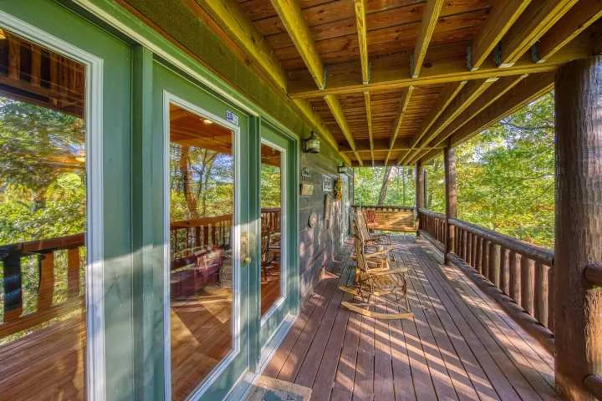 Two classic wooden rocking chairs are positioned on a deck with a rustic railing, facing out toward lush greenery and distant mountains. The inviting scene suggests quiet mornings, wildlife watching, and heartfelt conversations with friends or family.