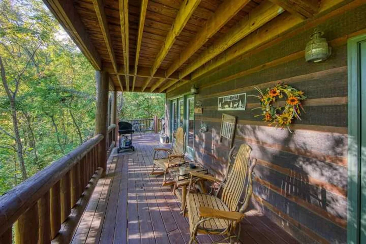 A close-up view of a wooden front porch with rocking chairs, a decorative wreath on the railing, and a grill in the background. The serene setting is framed by natural wood and glimpses of greenery, inviting guests to pause and soak in the ambiance before stepping inside.