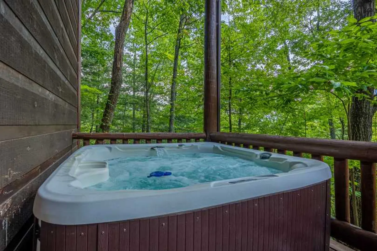 A serene wooden deck featuring a private hot tub surrounded by lush greenery. The vantage point suggests a peaceful spot for couples to relax, socialize, or stargaze under the clear mountain sky.