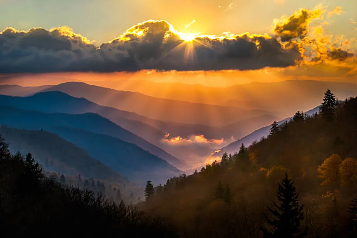 A breathtaking panoramic view of mountain ranges stretches into the distance, clouds streaked with sunset hues and valleys bathed in golden light. The scene captures the grandeur and serenity of the Smokies, inviting adventure and contemplation.