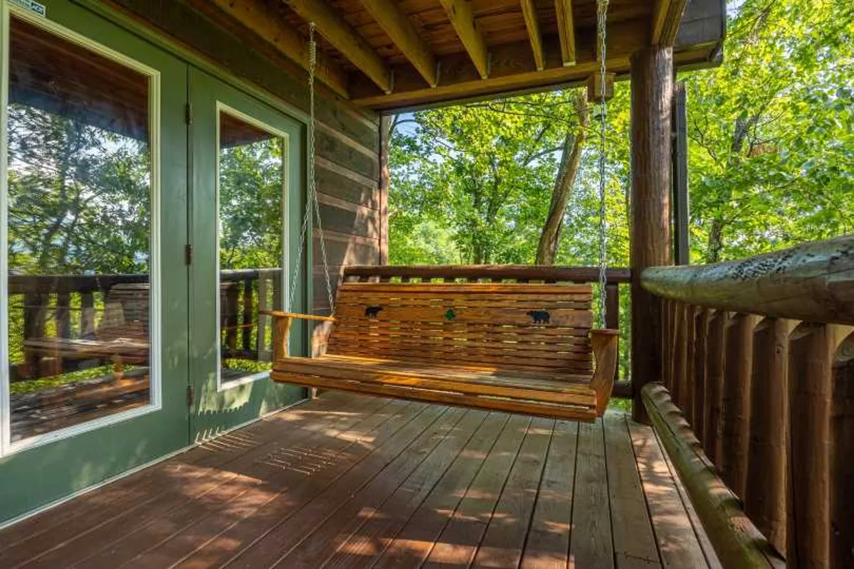 A cozy exterior scene featuring a wooden swing on a spacious porch, with large glass doors opening into the cabin and lush greenery surrounding the area. The atmosphere is serene and perfect for relaxation or family bonding after a day of outdoor activities.